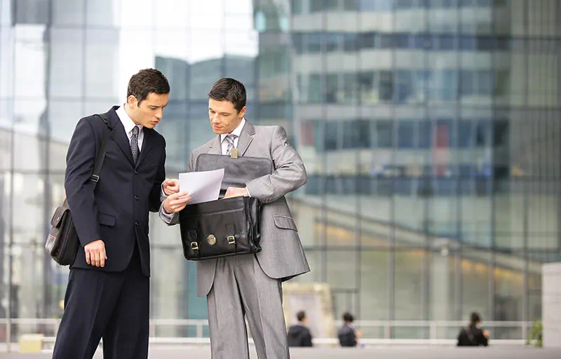 Two business men in suits outside of an office building look over some documents.